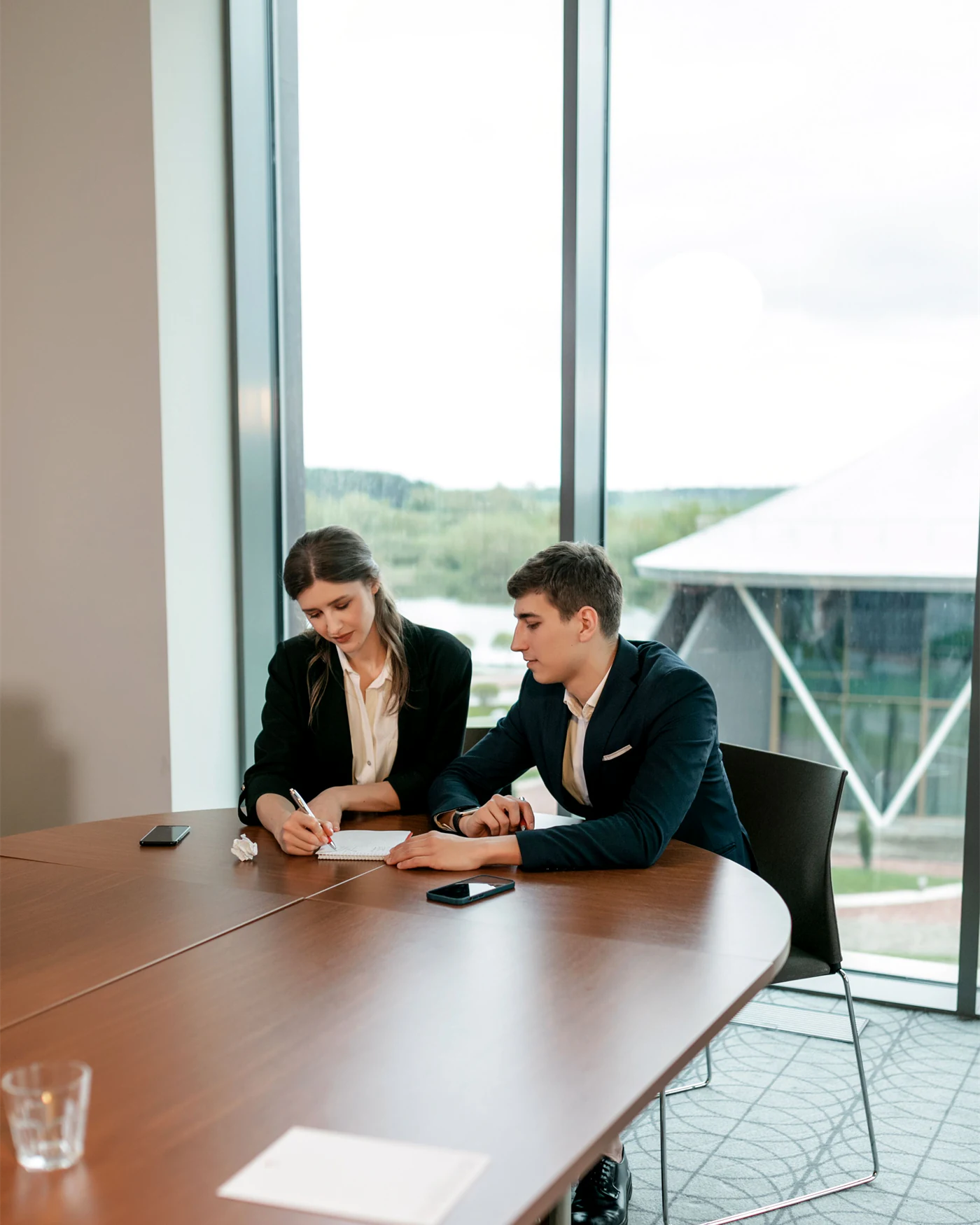 Two colleagues taking notes at a meeting table beside a large office window