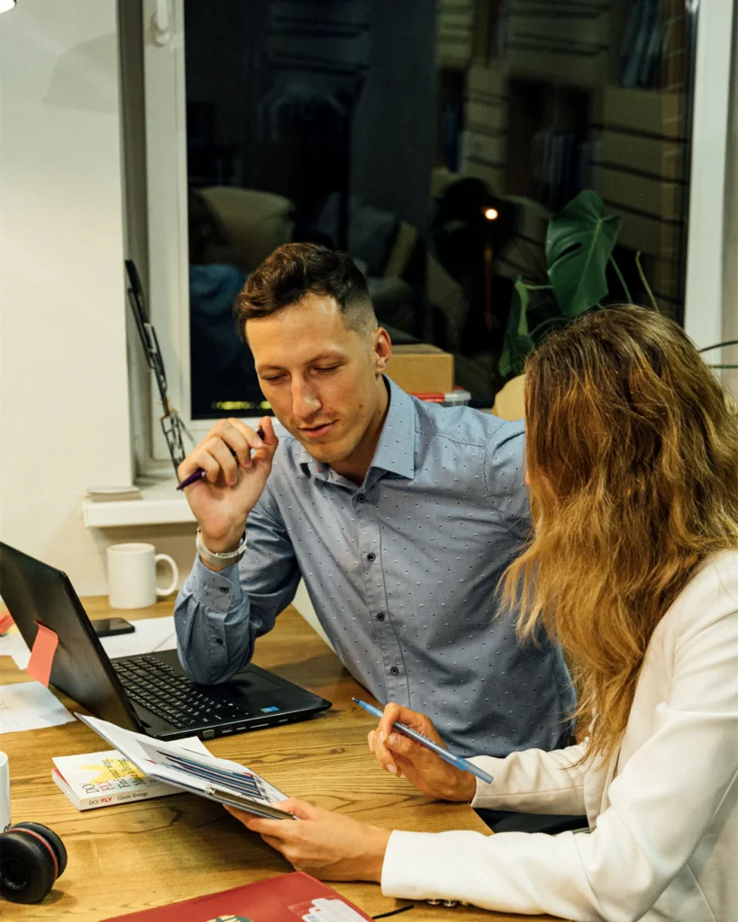 Two colleagues discussing plans spread across a wooden table