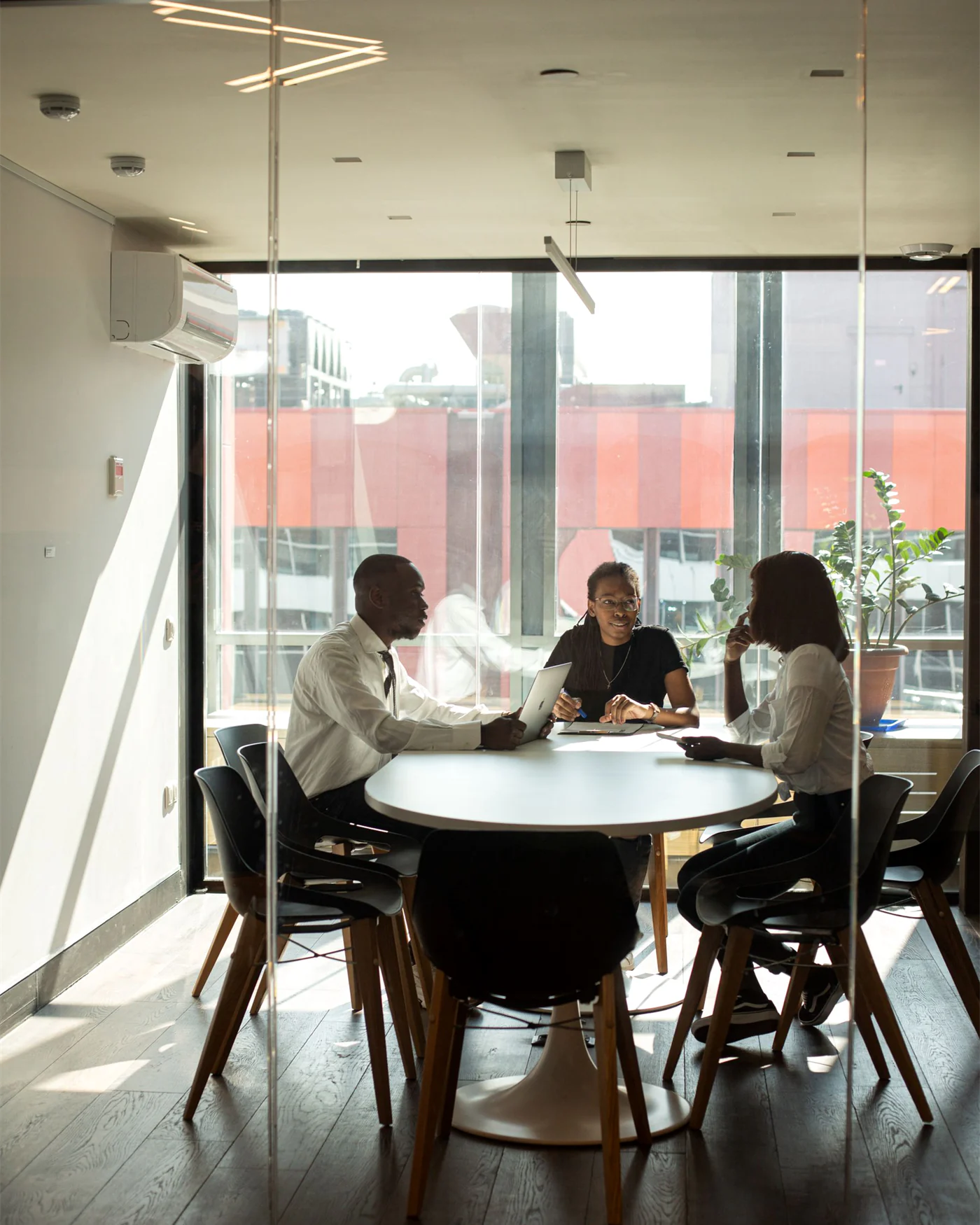 Three people meeting around a round table beside a large window