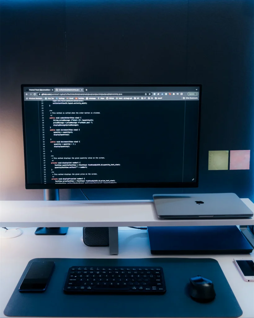 A monitor showing code on a clean desk with a laptop and keyboard