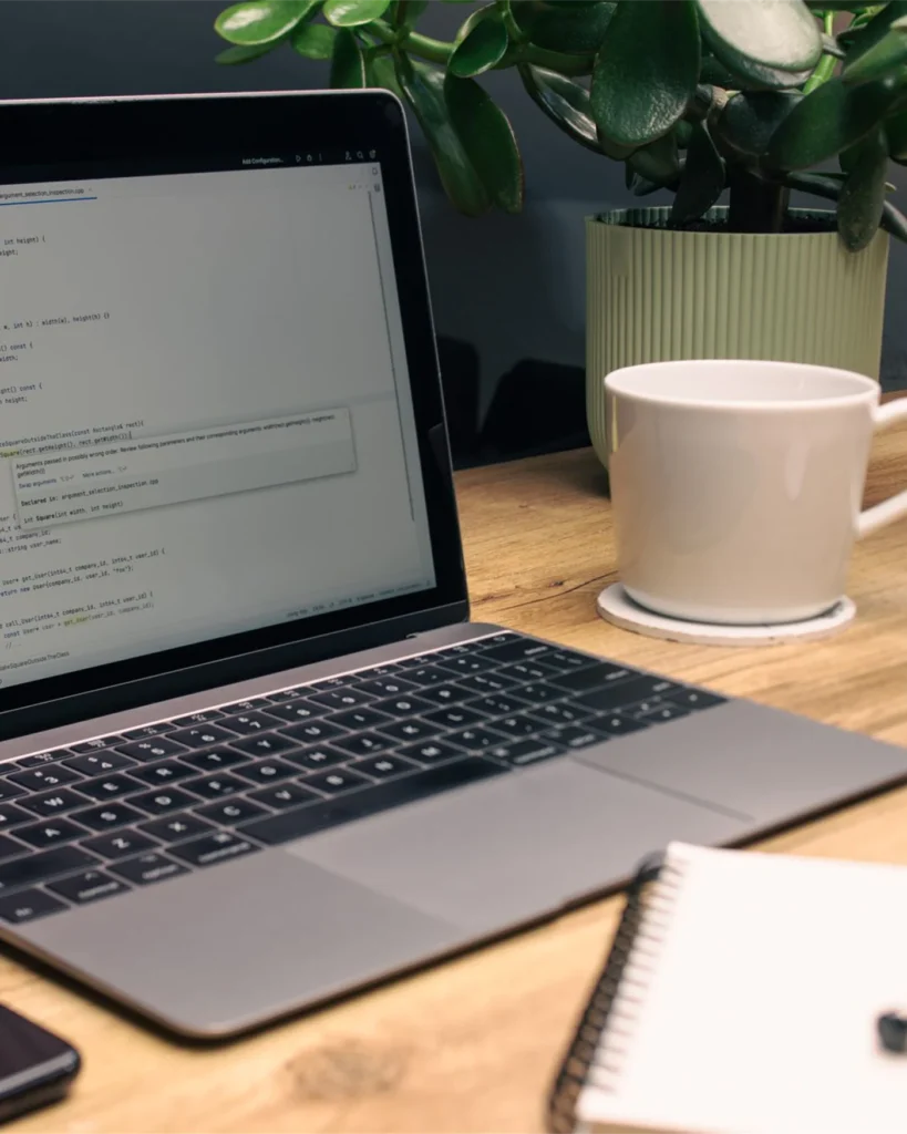 A laptop showing code on a wooden desk beside a notebook and coffee